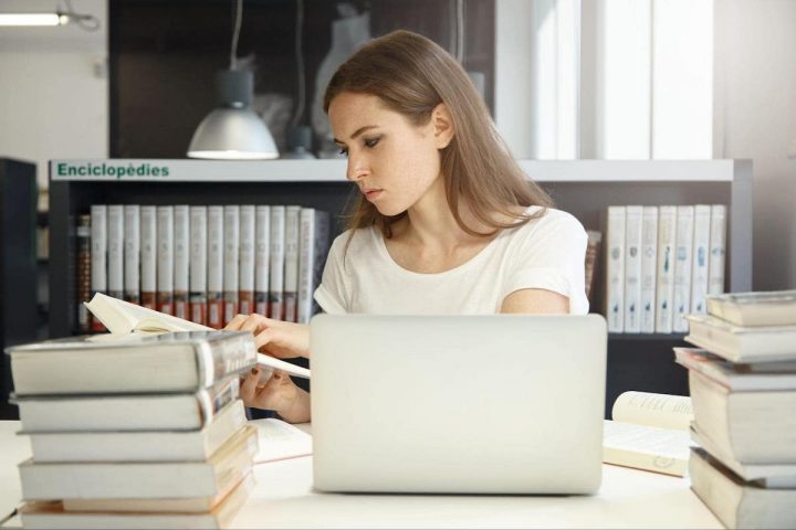 młoda kobieta siedząca przed laptopem i przeglądająca książkę